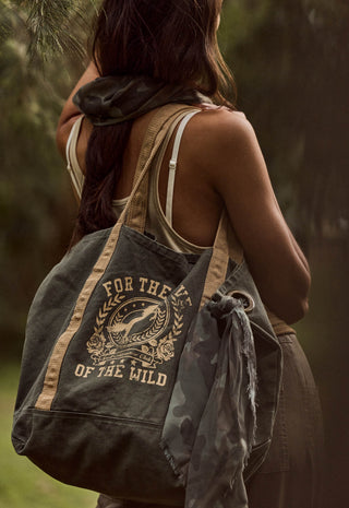 A woman outdoors carrying a large canvas tote bag over her shoulder. The bag is dark olive green with tan straps and a tan graphic print that reads 'FOR THE LOVE OF THE WILD' around a deer illustration.