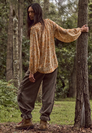A woman stands in a wooded outdoor setting, wearing a loose, long-sleeve patterned top in warm tan and brown tones, dark olive cargo pants, and brown hiking boots, with one hand resting on a tree trunk.