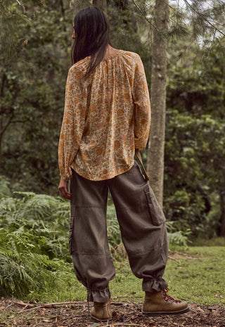 A person standing outdoors in a wooded area, shown from the back, wearing a loose long-sleeve blouse with an abstract print in warm tan, brown, and cream tones, paired with dark olive cargo pants and brown boots.