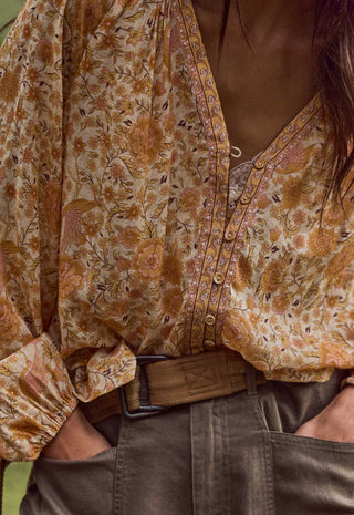 A close-up of a person wearing a loose, long-sleeve button-front blouse with a warm floral and paisley print in tan, rust, and brown tones over a cream base, tucked into dark olive pants with a brown belt.