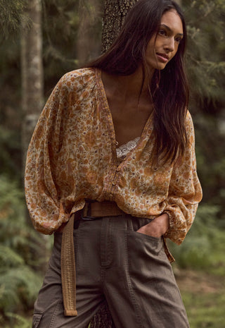 A woman standing outdoors in a forest wearing a loose, long-sleeve boho-style blouse with an abstract floral pattern in tan, brown, and muted orange, paired with dark olive cargo pants and a woven belt.
