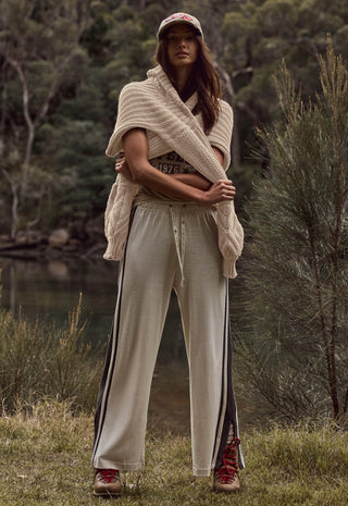 A woman stands outdoors in a wooded setting wearing a beige knit wrap, beige wide-leg pants with dark side stripes, a baseball cap, and red sneakers.