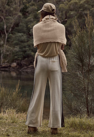 A person standing outdoors by a body of water, shown from the back, wearing a beige knit shrug, beige top, beige wide-leg pants with dark side stripes, and a beige cap.