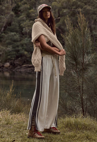A woman stands outdoors near a body of water and trees, wearing a chunky knit beige wrap or poncho over cream wide-leg pants with dark side stripes, a beige cap, and brown lace-up boots.