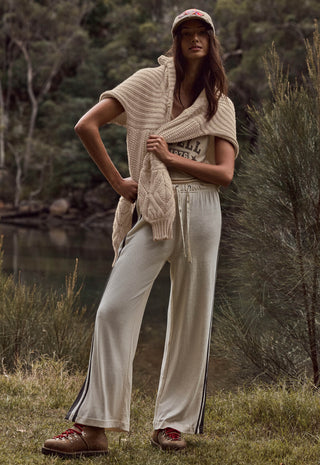 A woman stands outdoors near a body of water and trees, wearing a cream knit cardigan draped over her shoulders, cream wide-leg pants with dark side stripes, a graphic T-shirt, brown clogs, and a pink baseball cap.